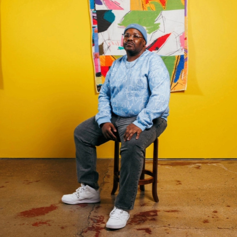 Color photograph of Herron alum Julian Jamaal Jones seated on a stool in front of one of his colorful quilts on display at his show at the Tube Factory ArtSpace.