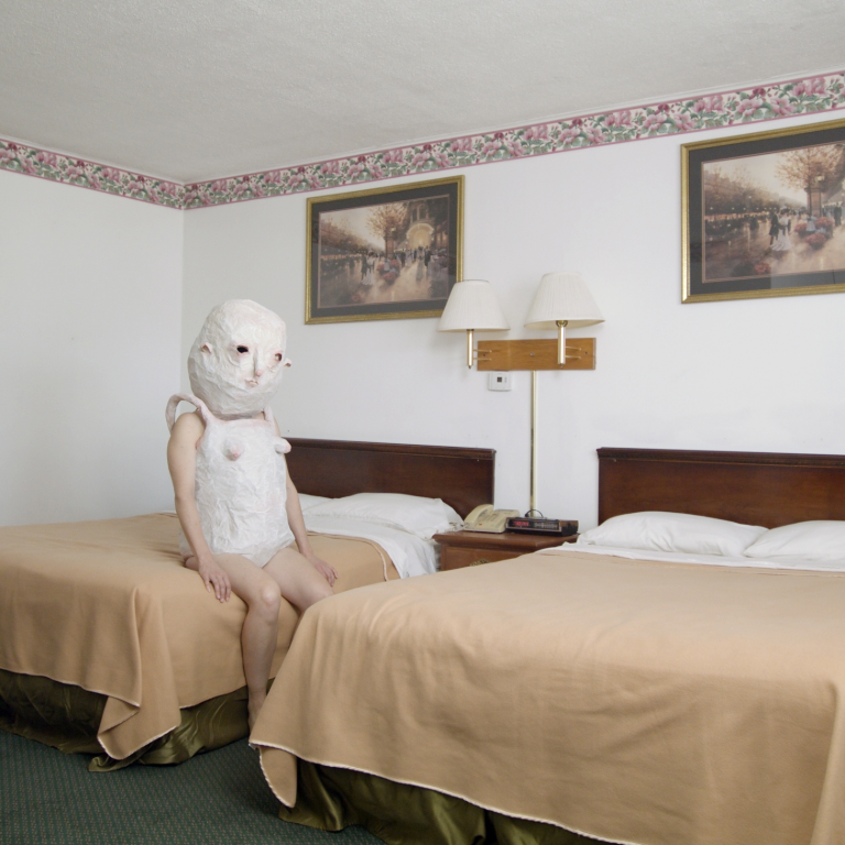 Still from artist Johanna Winters' film HOWW TO WAYT featuring a white person with a papier mache costume seated on a motel bed.